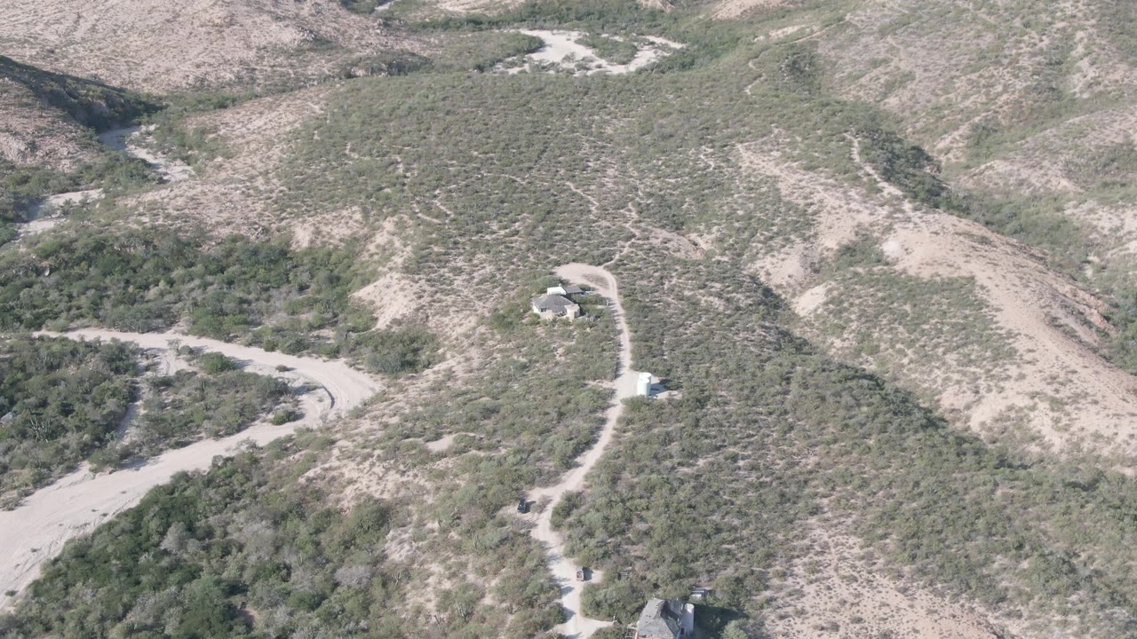 Mid-approach aerial frame in Cabo Pulmo showing a dirt road bending across a shrub-covered slope toward small structures, with dry hills beyond and no visible people.