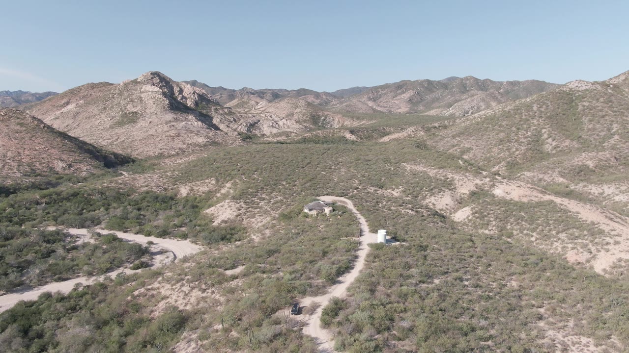 Aerial view in Cabo Pulmo, Baja California Sur, with a pale dirt road curving through scrub-covered hills toward rocky mountains under a clear sky, no visible people.
