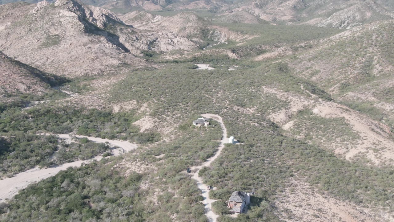 A high aerial frame over Cabo Pulmo, Baja California Sur shows a winding dusty road below rocky shrub-covered hills, a small structure near the lower edge, and no visible people.