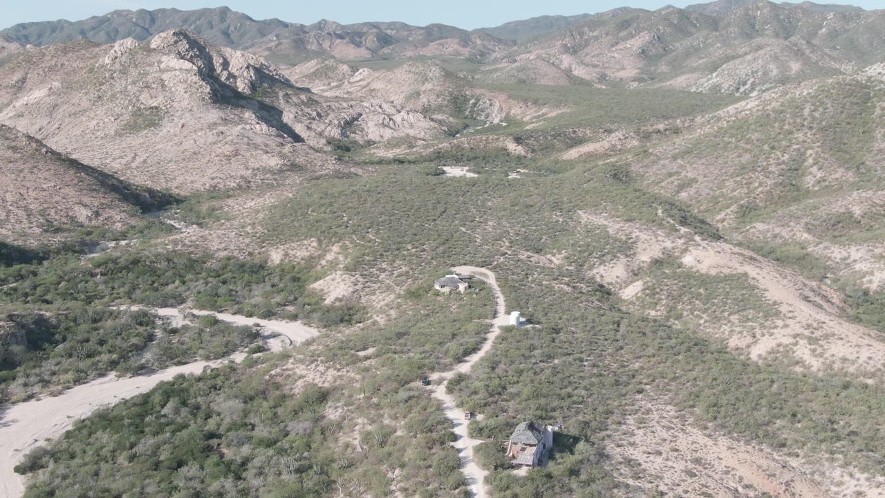 At 0:01, the Cabo Pulmo aerial view keeps the dusty road and small structure low in frame while pale ridges and scrubby slopes fill the background, with no visible people.