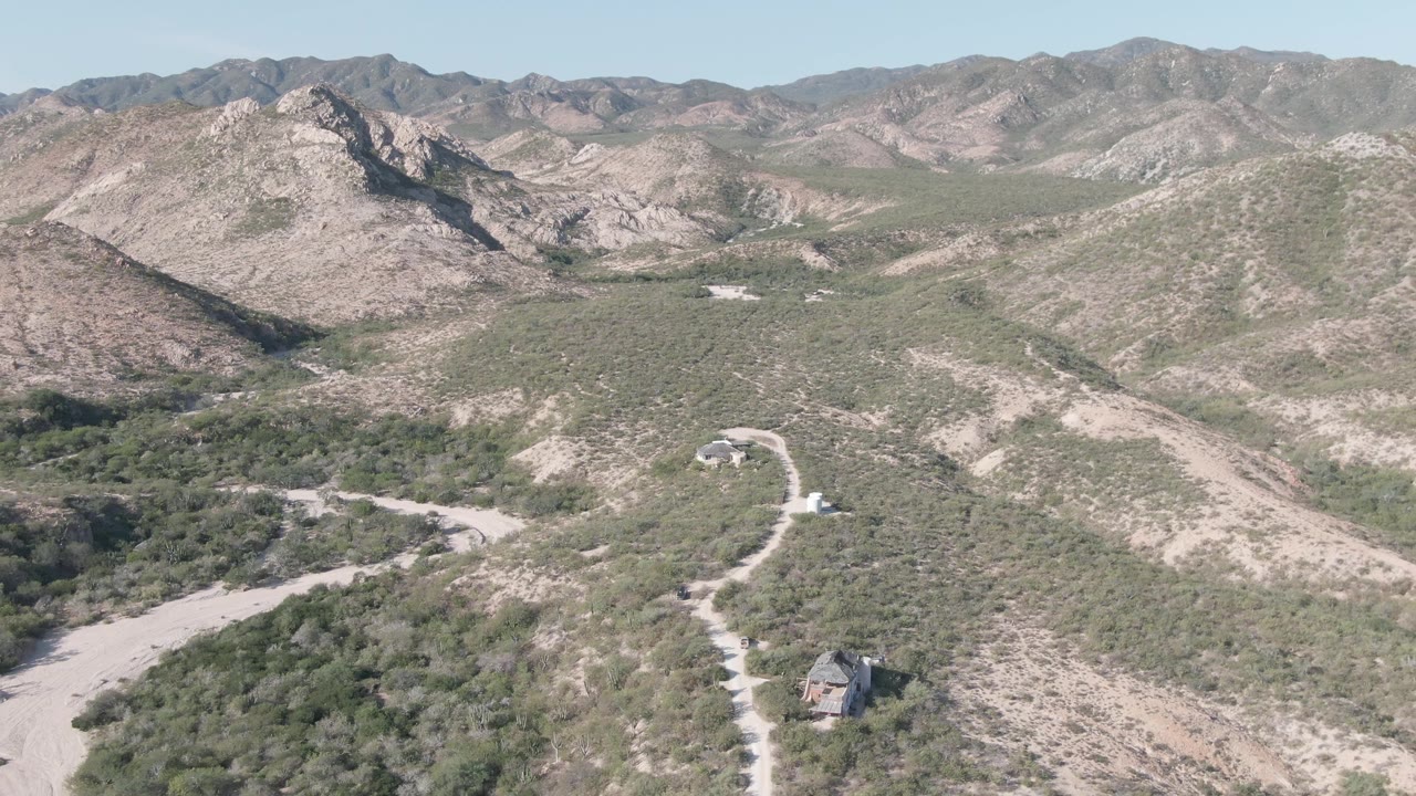 At 0:02, the drone perspective shows the road bending through scrubland toward layered rocky hills in Cabo Pulmo, with the isolated building still visible and no visible people.