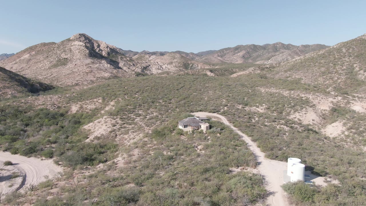 Opening aerial frame over Cabo Pulmo desert terrain with a small round-roofed house near a dirt road, a white water tank at the lower right, distant rocky mountains, and no visible people.