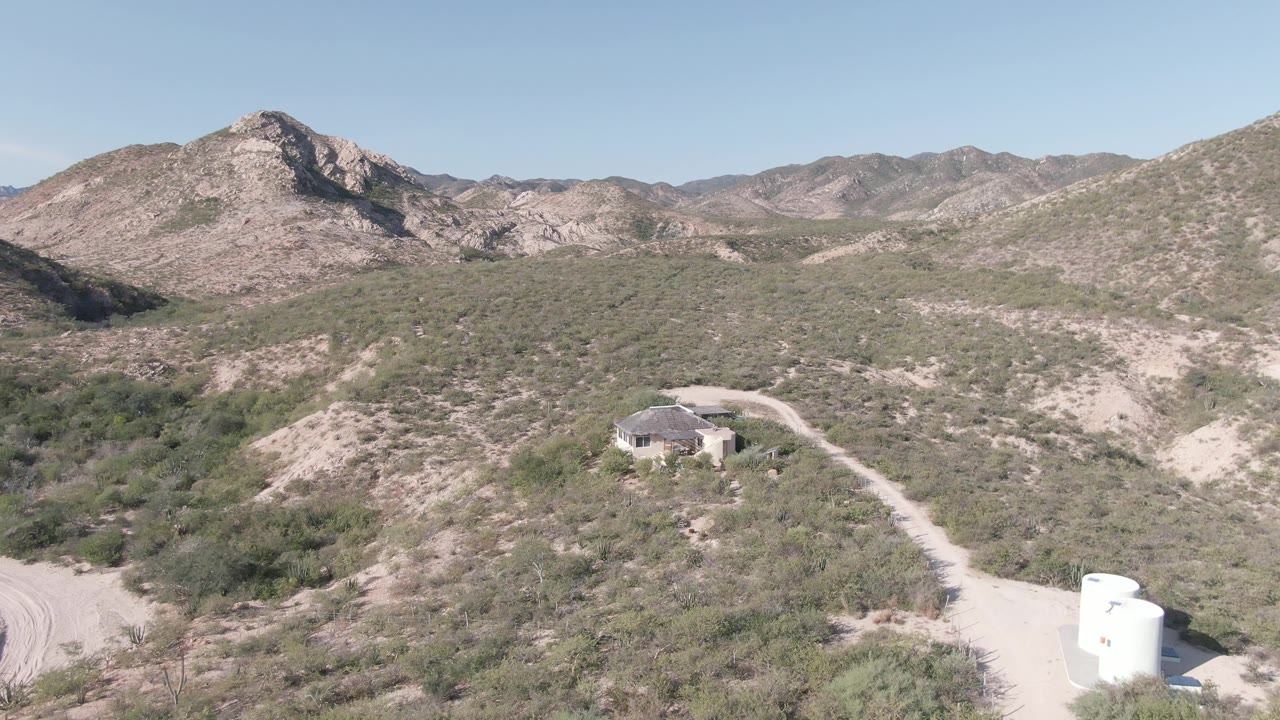 Aerial view in Cabo Pulmo with the small desert house slightly closer, pale dirt road curving through scrub vegetation, rocky mountain ridges in back, and no visible people.