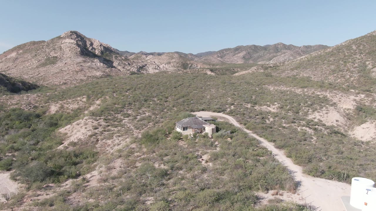 Cabo Pulmo aerial frame showing the house and access road below scrub-covered hills, the white water tank now near the far lower right edge, with no visible people.