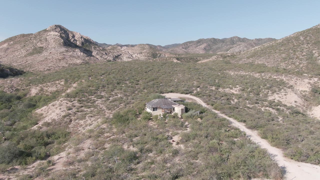Wide aerial desert landscape with a house, winding dirt road, and mountains.