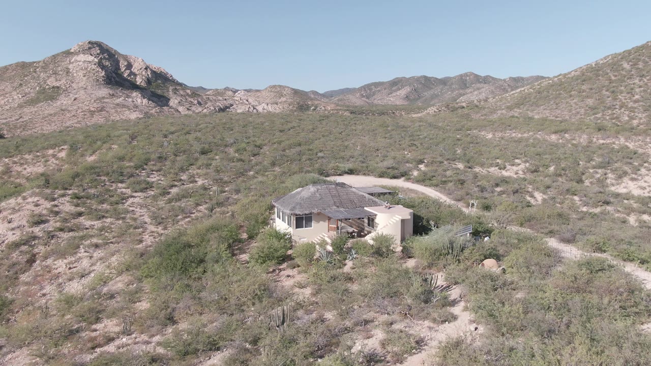 Opening frame of a pale house in Cabo Pulmo, Baja California Sur surrounded by desert scrub, with a dirt road curving behind it, rocky mountains in the background, and no visible people.