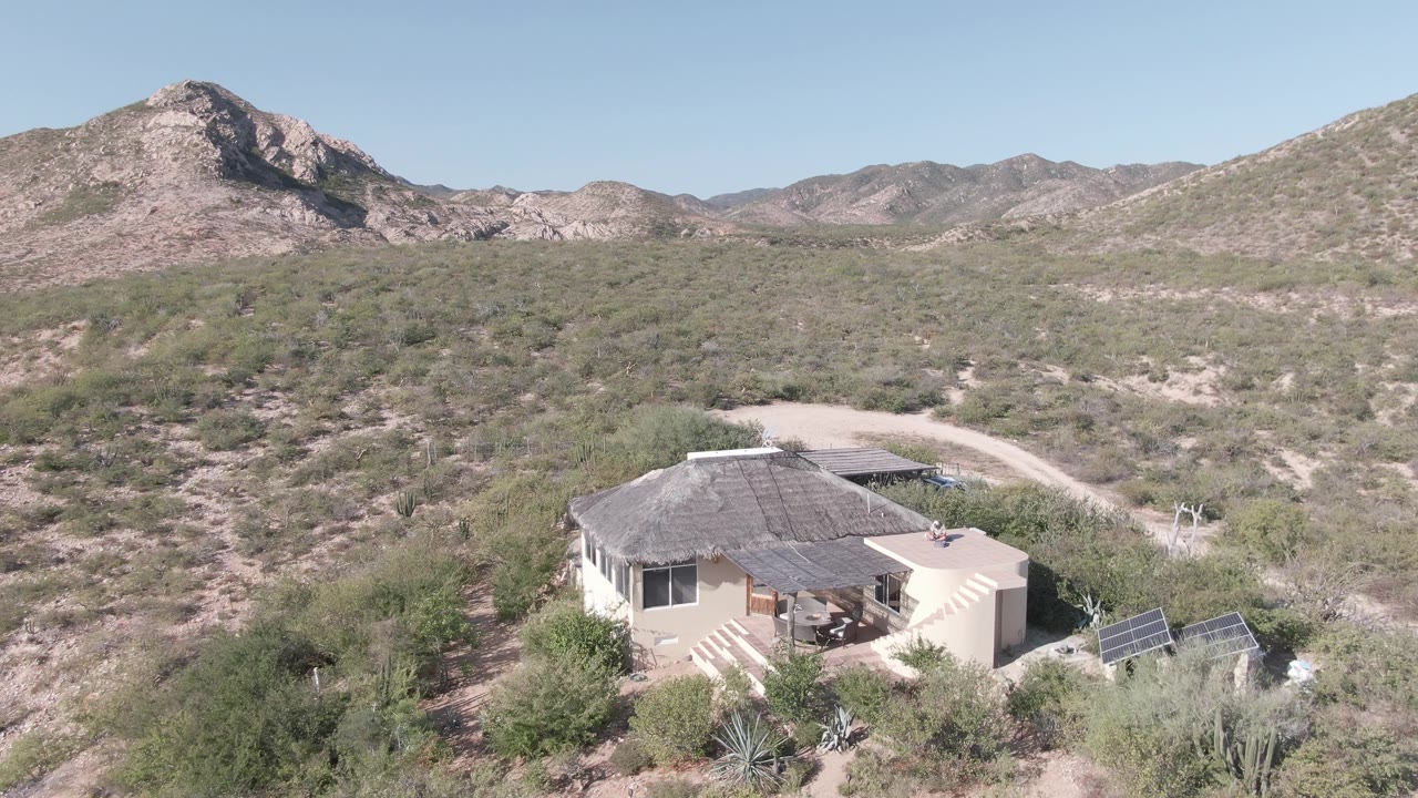 Cabo Pulmo desert frame with the house set low right of center, solar panels beside the structure, a pale dirt road behind it, dry scrub throughout the foreground, and no visible people.