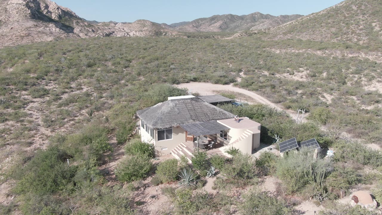 Aerial view of a pale house with solar panels, scrub vegetation, a dirt road, and distant mountains in Cabo Pulmo, Mexico, with no visible people.