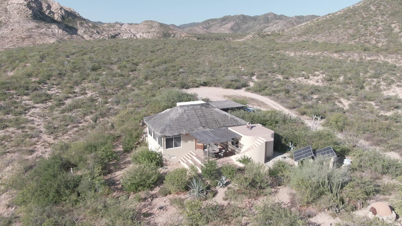 The house sits left of center with solar panels to the right, low desert shrubs around it, and a curving dirt road behind it in Cabo Pulmo, with no visible people.