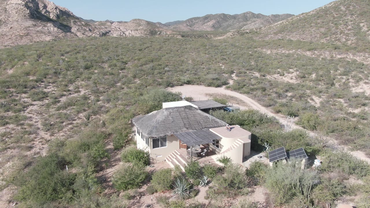 A wider aerial frame places the solar-equipped house below rocky hills and dense desert scrub in Cabo Pulmo, Mexico, with no visible people.