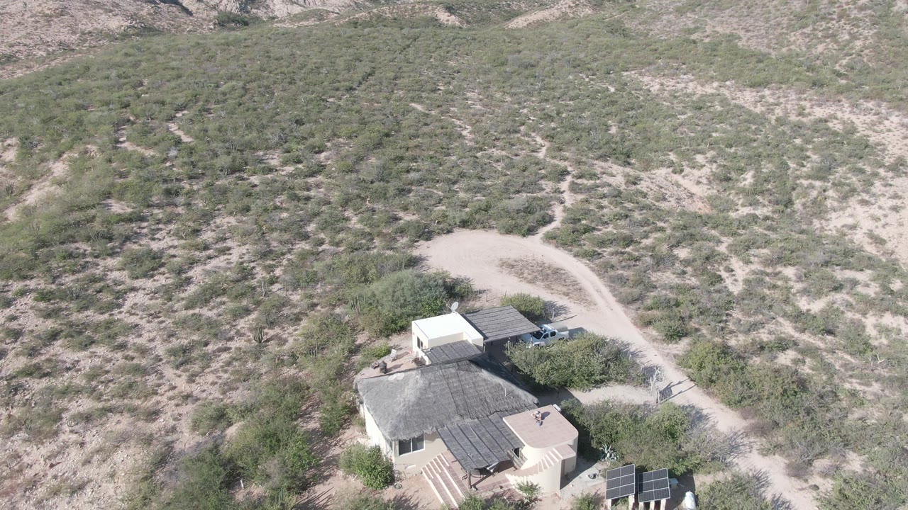 High aerial frame over a house in Cabo Pulmo, Mexico, with a pale dirt road curving behind it, sparse shrubs on sandy hillsides, solar panels near the lower right, and no visible people.