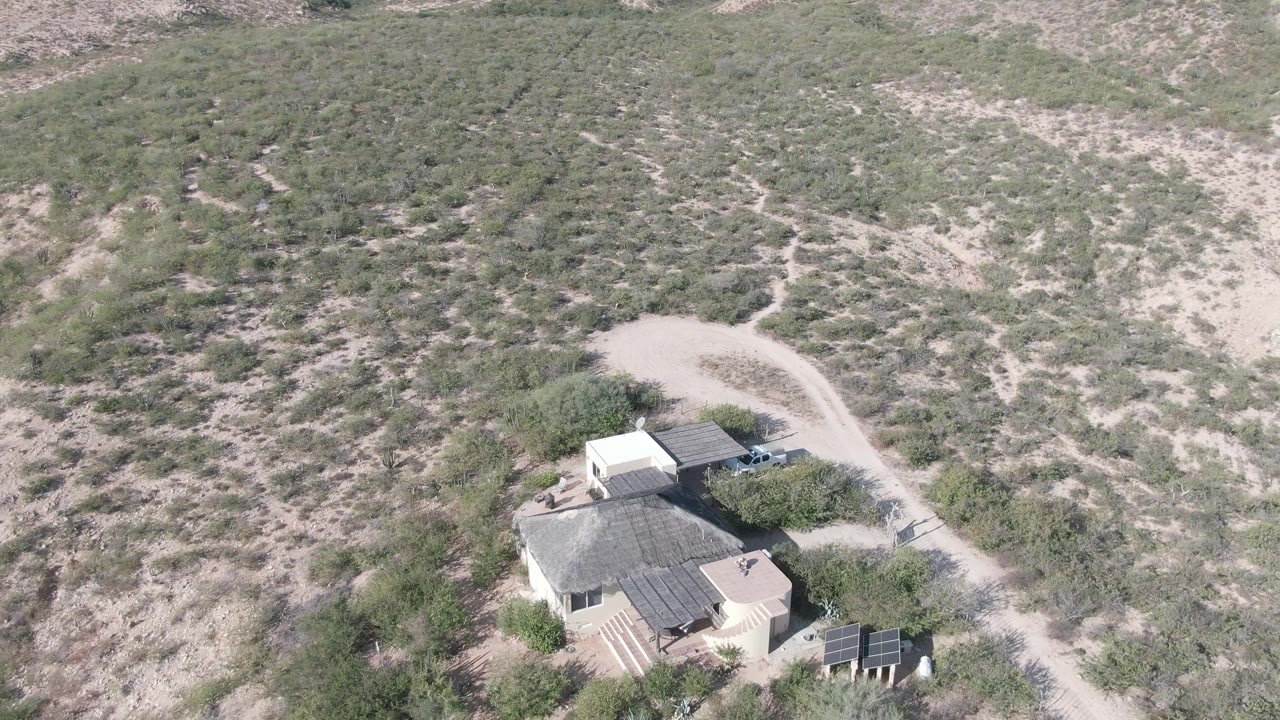 Desert house and nearby vehicle seen from above amid sandy ground and shrubs.