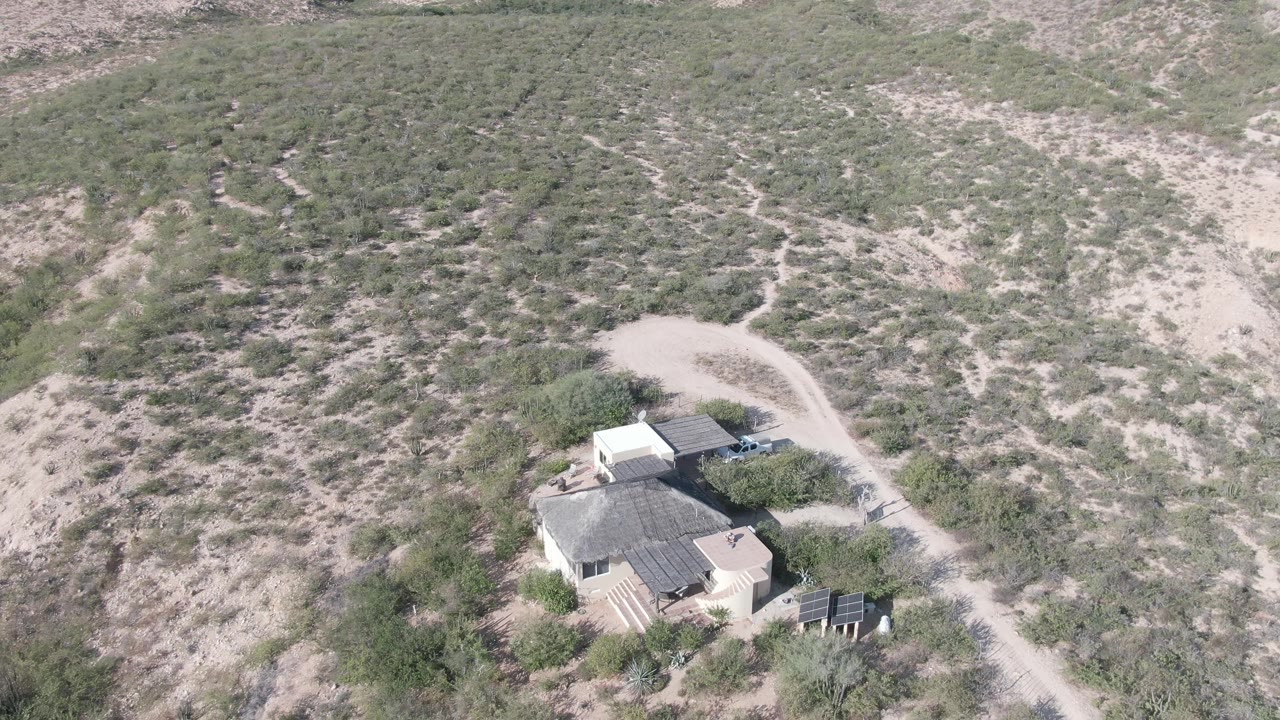 High oblique frame over a Cabo Pulmo desert house, with solar panels visible near the lower right, a light dirt road behind the buildings, scrubby hillsides, and no visible people.
