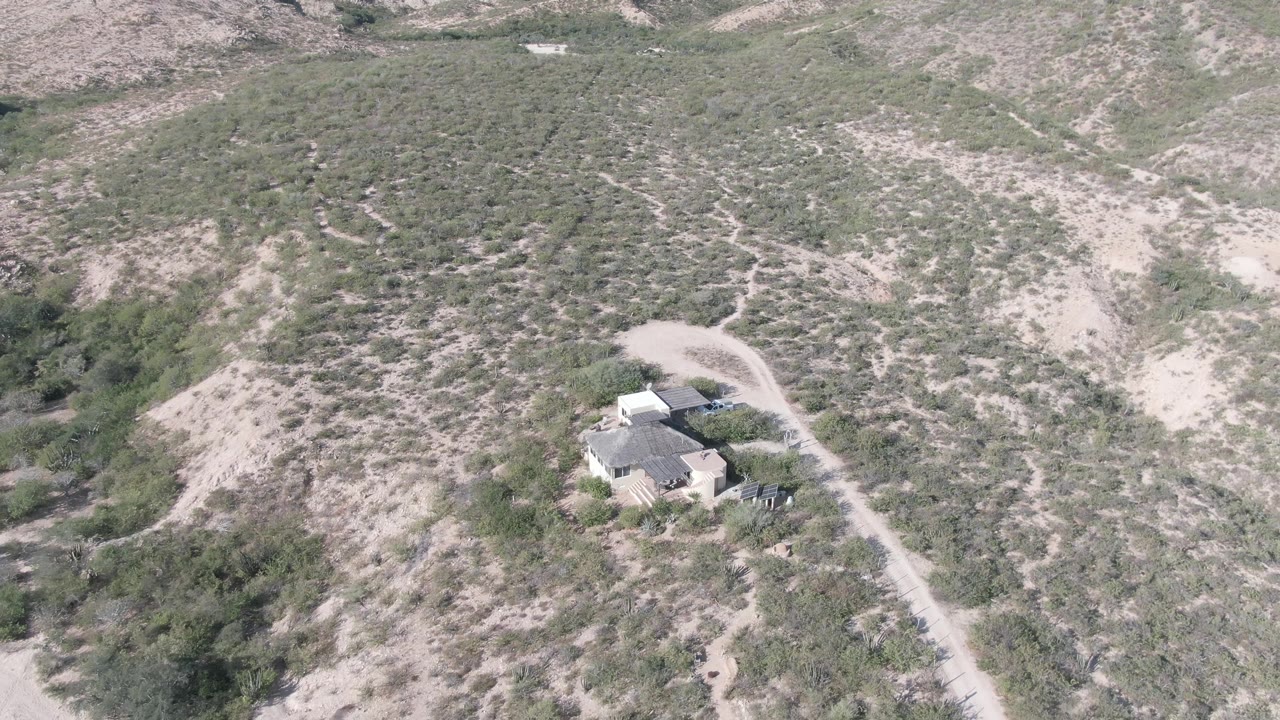 Aerial frame over Cabo Pulmo desert hills shows an isolated house near a pale dirt road, scrub vegetation, rocky slopes, and no visible people.
