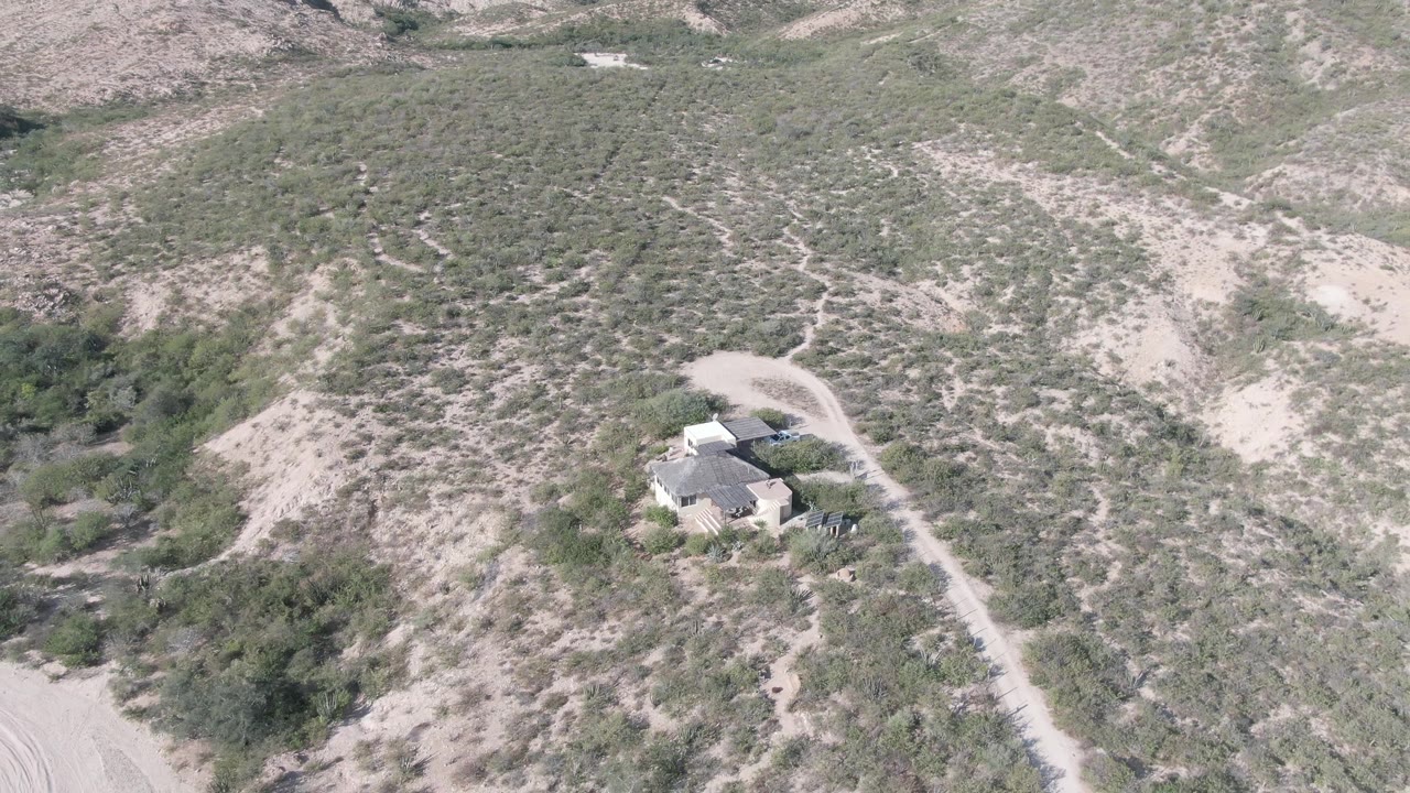 Cabo Pulmo aerial view keeps the house and dirt road in the midground while dry scrub, pale soil, and ridged hills fill the frame, with no visible people.