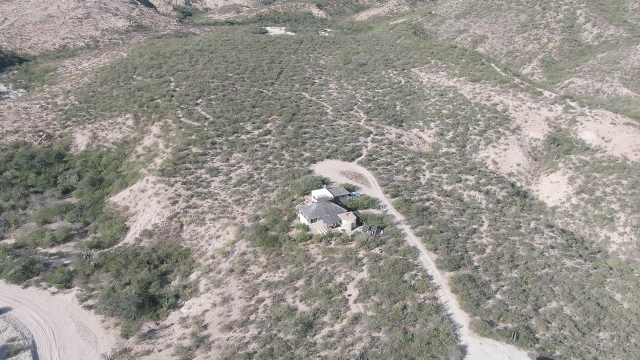 The Cabo Pulmo frame shows the house set off a curving dirt road, with brushy slopes, dry gullies, and no visible people in the open terrain.