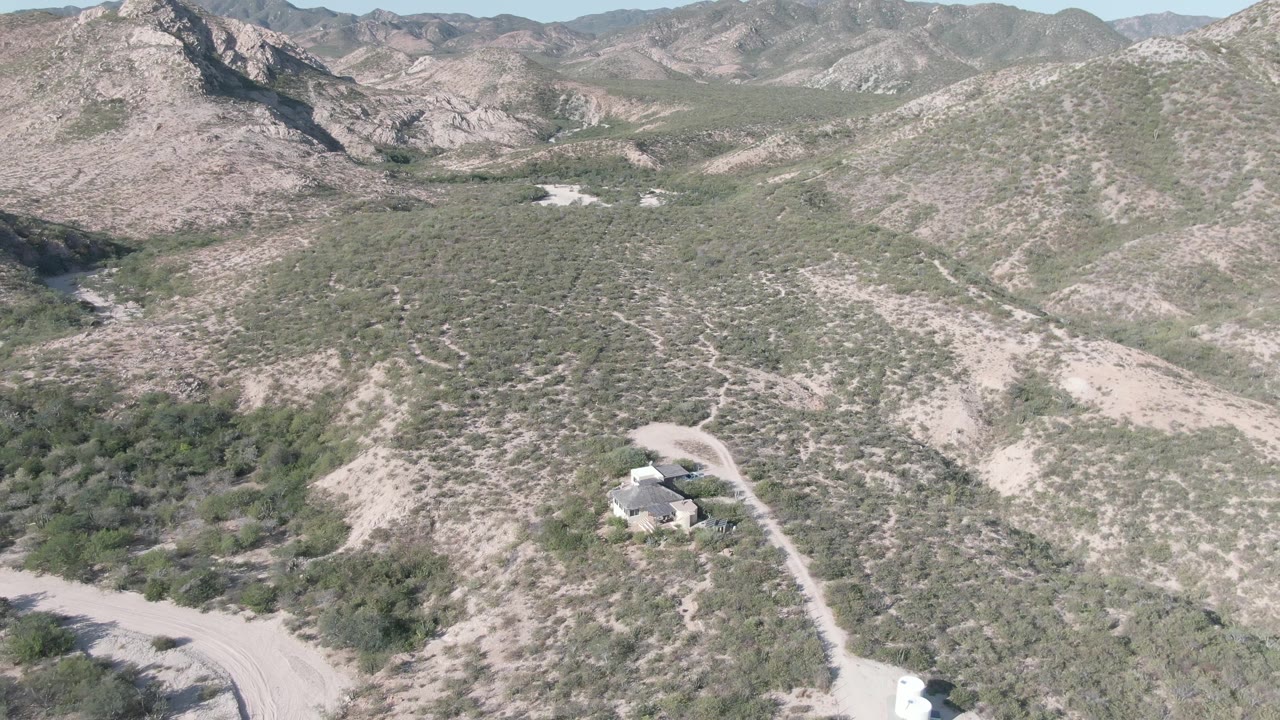 Aerial frame over Cabo Pulmo desert hills with a winding dirt road at the lower left, a small house on the slope, layered mountains in back, and no visible people.