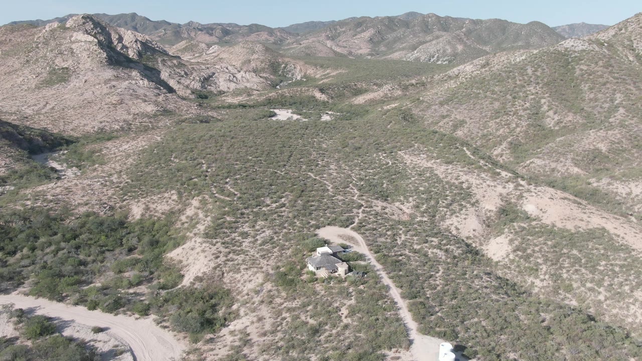 Wide aerial view in Cabo Pulmo showing pale rocky slopes, patchy desert shrubs, a small house near a curving dirt road, distant ridgelines, and no visible people.
