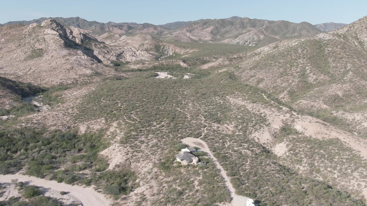 Cabo Pulmo aerial frame with the winding road and isolated house near the lower right, scrub-covered hills across the center, rocky mountains beyond, and no visible people.