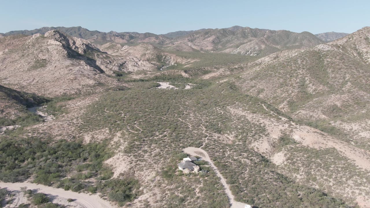 Desert mountain panorama with sparse vegetation, dirt tracks, and a house on a slope.