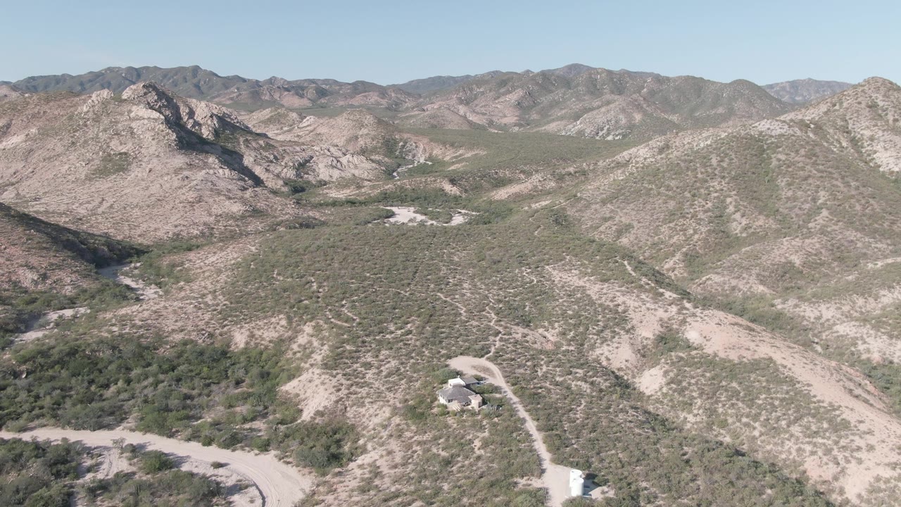Aerial view near Cabo Pulmo, Baja California Sur, showing scrub-covered tan hills, a pale dirt road in the lower frame, distant mountain ridges, and no visible people.