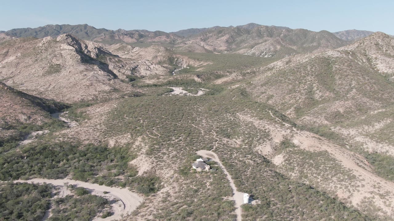 High aerial view near Cabo Pulmo, Baja California Sur, showing pale rocky slopes, scattered green vegetation, a dirt road near the bottom edge, distant ridges, and no visible people.