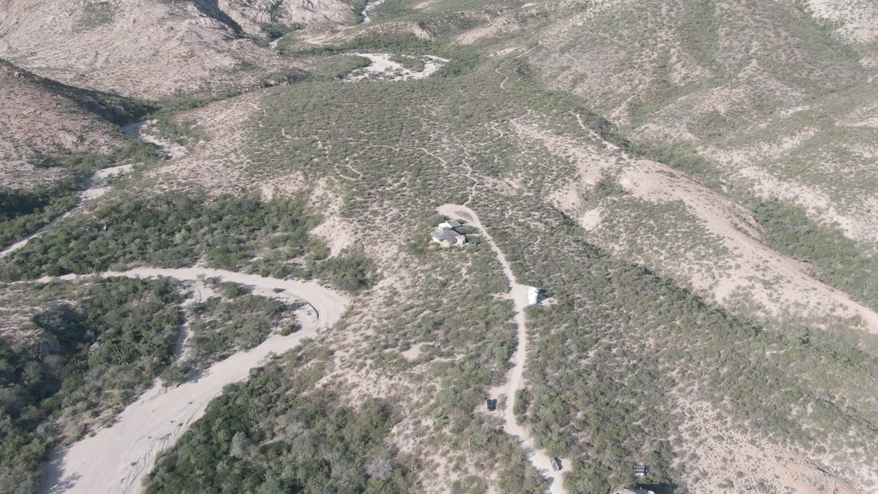 Aerial view in Cabo Pulmo, Baja California Sur, centered on a curved dirt road below scrub-covered slopes and a small pale structure, with no visible people.