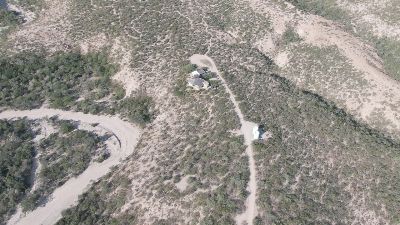 Aerial frame over Cabo Pulmo, Baja California Sur, showing pale dirt roads, scrub-covered hills, and small light-roofed structures with no visible people.