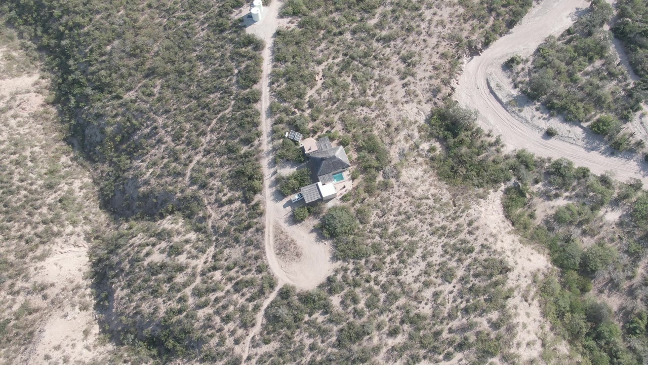 Overhead frame from Cabo Pulmo, Baja California Sur, showing a vehicle on a pale dirt track near a small house, scrub vegetation, sandy ground, and no visible people.