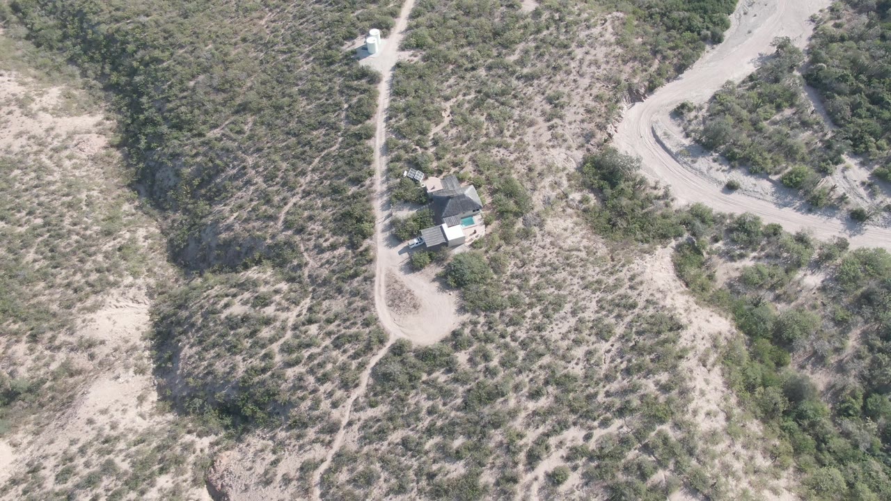 Aerial frame over Cabo Pulmo, Baja California Sur, showing a small hillside house near a pale dirt road, scrub-covered slopes, sandy ground, and no visible people.