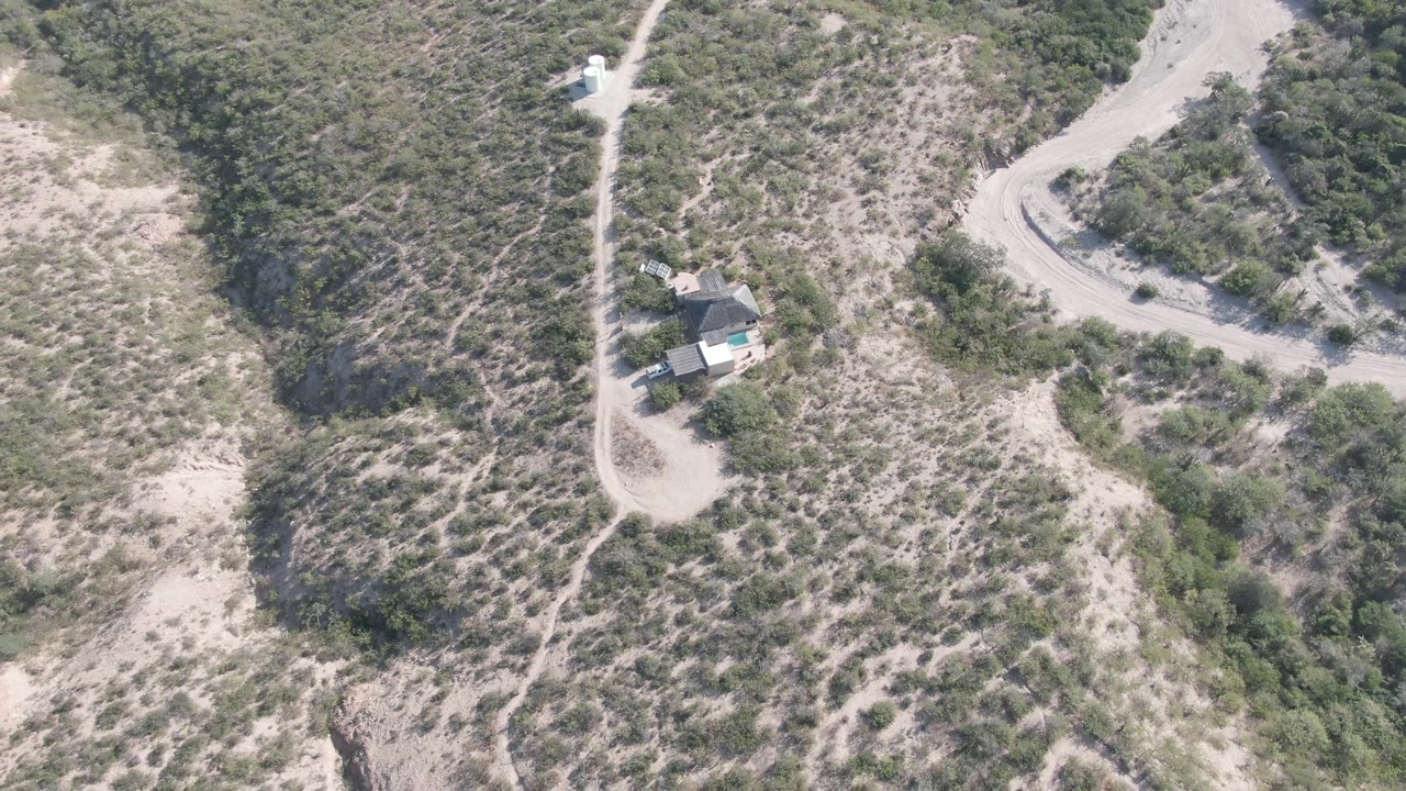 High aerial view in Cabo Pulmo with the small house lower in frame, a winding dirt road curving through pale sand, dense brush patches, and no visible people.