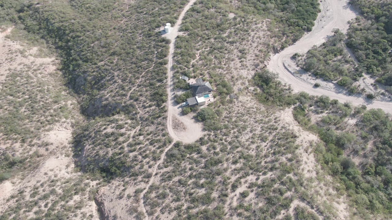 Aerial frame over Cabo Pulmo showing the house near the lower left, a narrow path running up the hillside, dry brush, pale soil, and no visible people.