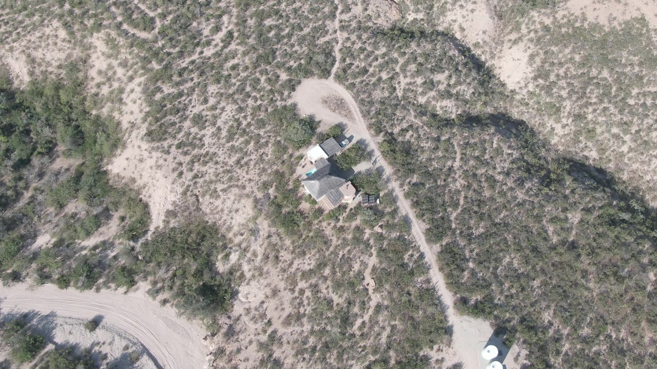 High aerial frame over Cabo Pulmo desert terrain with a small roofed structure beside a pale dirt road, sparse scrub, sandy ground, and no visible people.