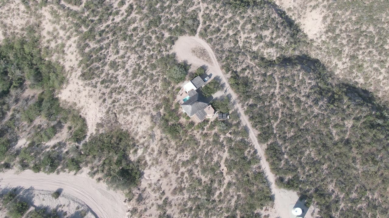 Cabo Pulmo aerial frame with a small building cluster near the dirt road, pale sand, darker scrub patches, and no visible people.