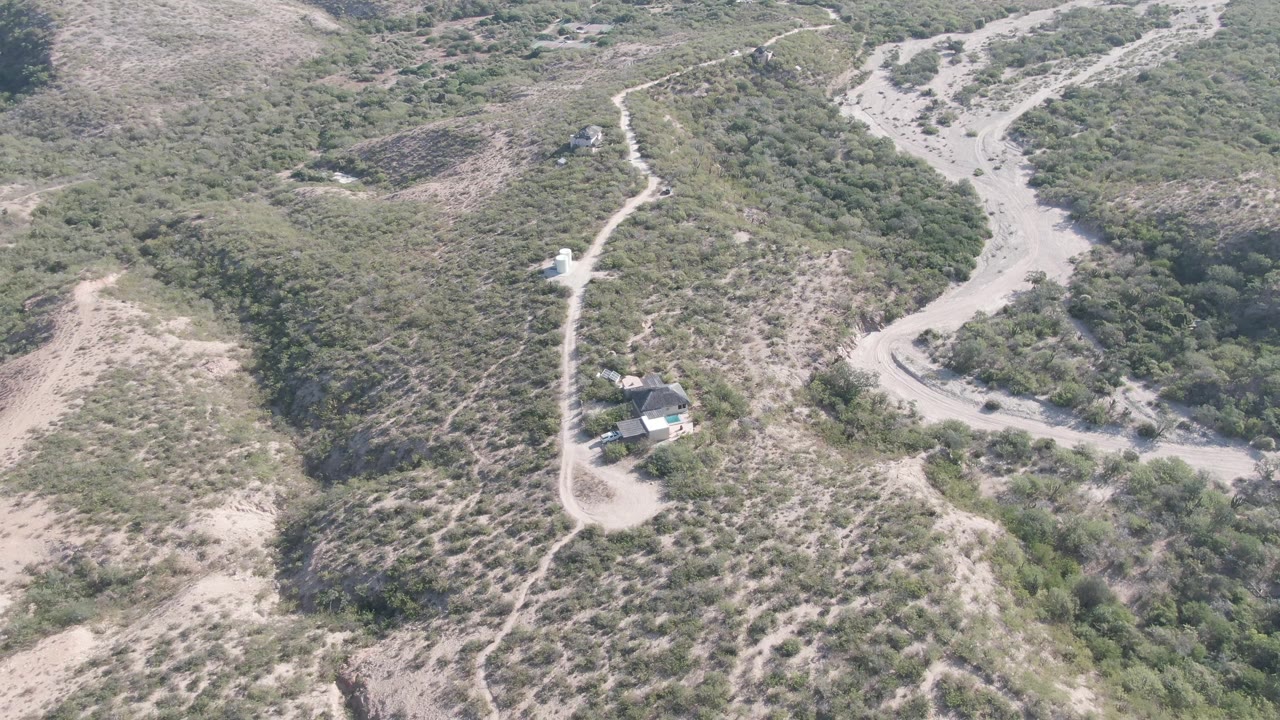 Aerial frame over Cabo Pulmo, Baja California Sur showing dry green-brown hills, pale winding dirt paths, a small roadside structure, and no visible people.