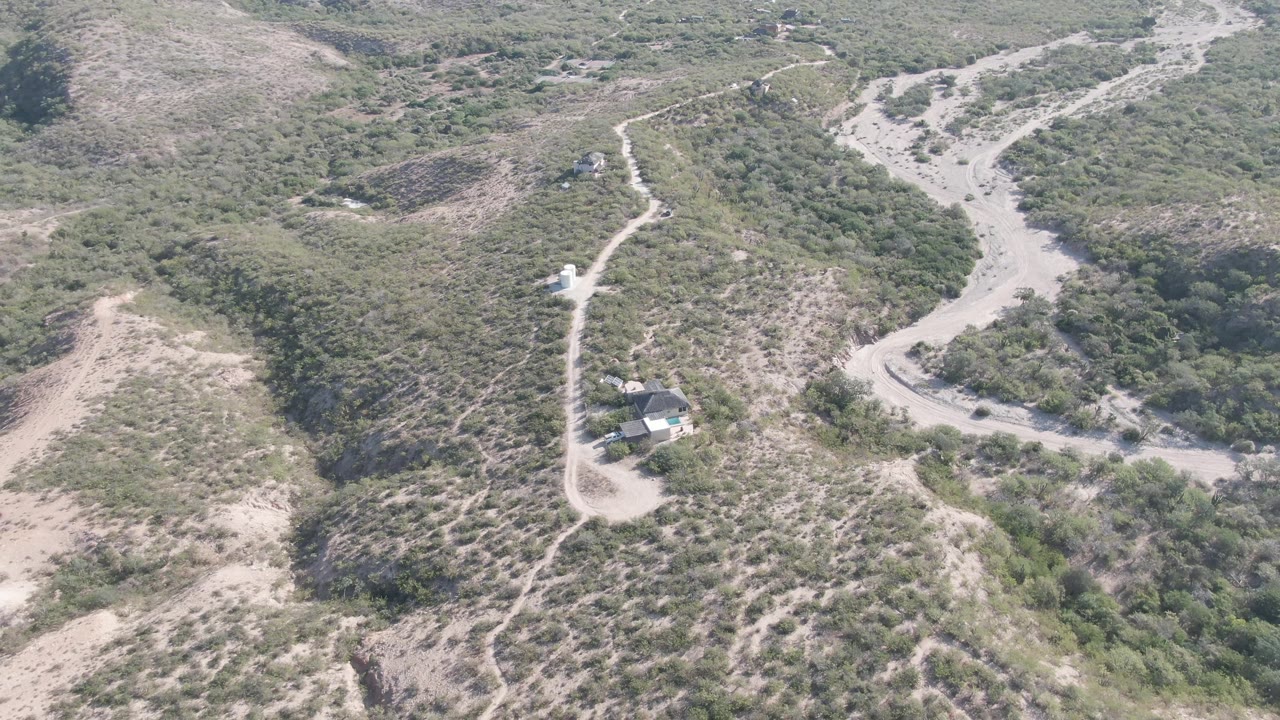 Aerial view in Cabo Pulmo, Baja California Sur with a pale dirt road curving through scrubby hills and the small structure still visible near the lower edge, with no visible people.