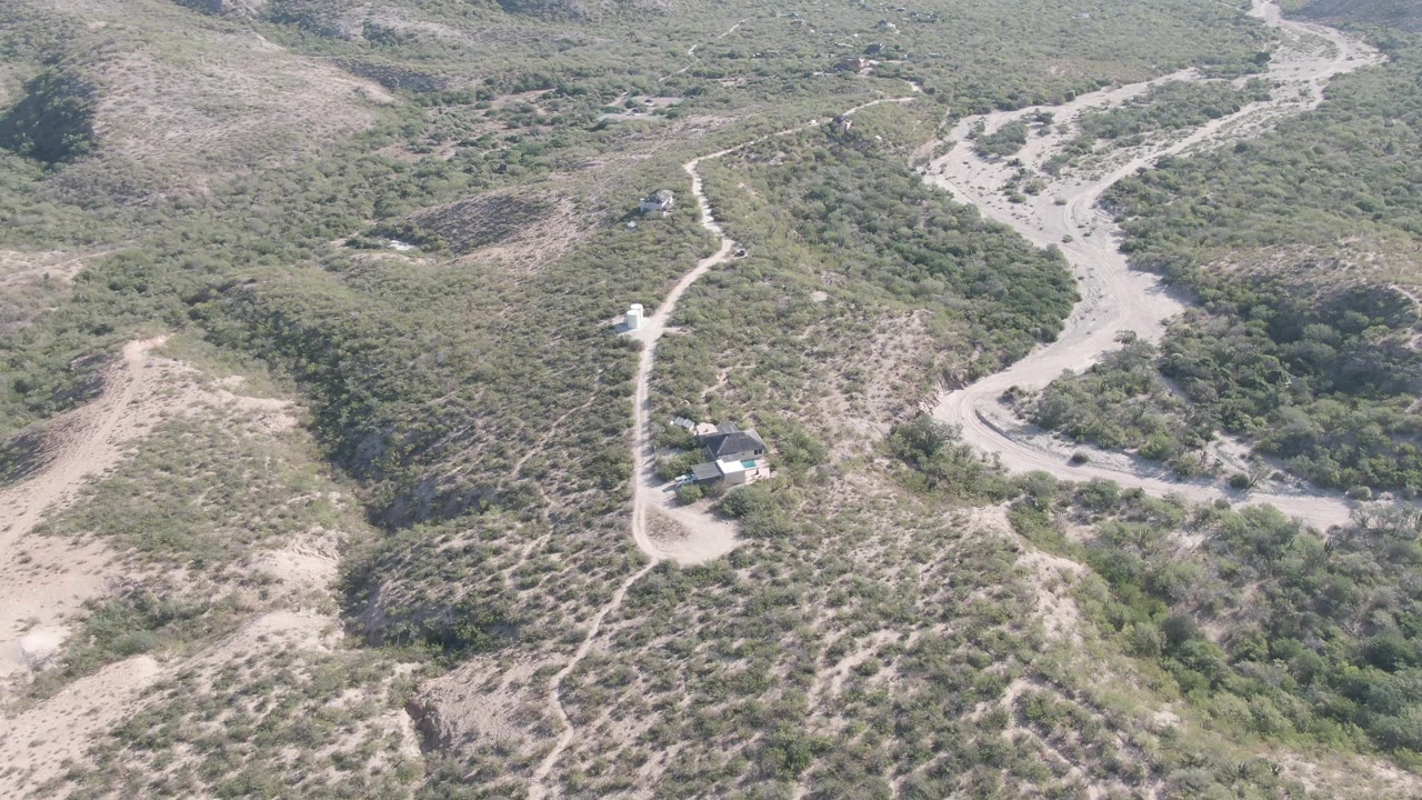 Cabo Pulmo aerial frame showing a dry wash or road cutting through green-brown hillside scrub, with a small structure and vehicle below and no visible people.