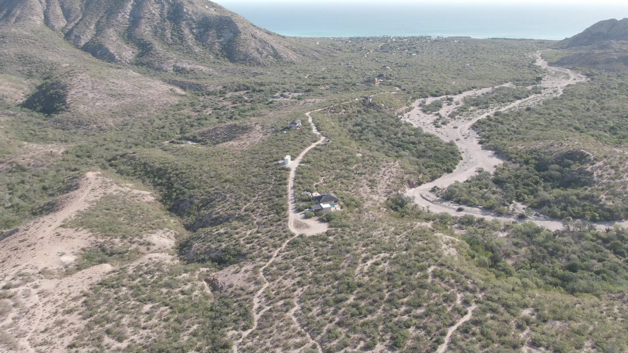 Aerial frame over Cabo Pulmo, Baja California Sur, with a winding dirt road, scrub-covered hills, rugged mountains, distant ocean, small rural structures, and no visible people.