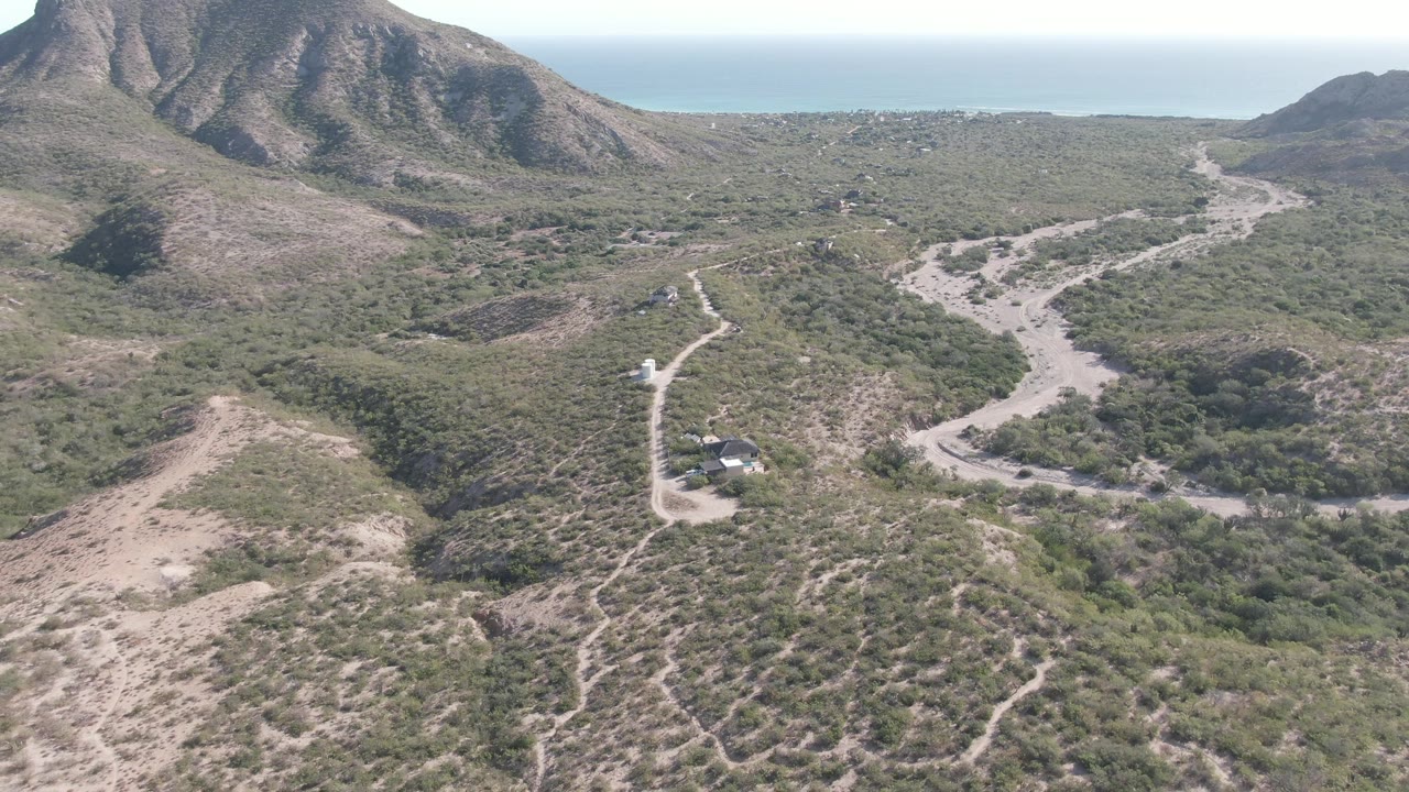 Aerial view in Cabo Pulmo showing the vehicle farther along the pale dirt road, with green-brown hills, mountain slopes, distant ocean horizon, and no visible people.