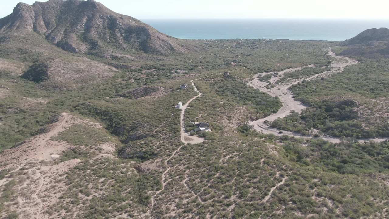 Cabo Pulmo aerial frame with the dirt road curving through scrubland, a small vehicle near the lower road, dark mountain faces in back, ocean on the horizon, and no visible people.