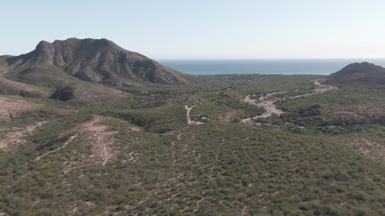 Opening frame over Cabo Pulmo coastal terrain with scrubby hills, a winding pale road, rugged mountains, blue ocean on the horizon, and no visible people.