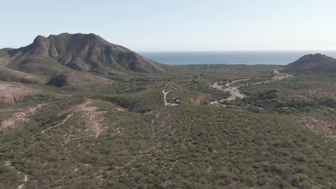 Cabo Pulmo aerial frame with the central mountain mass more prominent, road bends visible through the scrubland, a smaller hill to the right, ocean beyond, and no visible people.