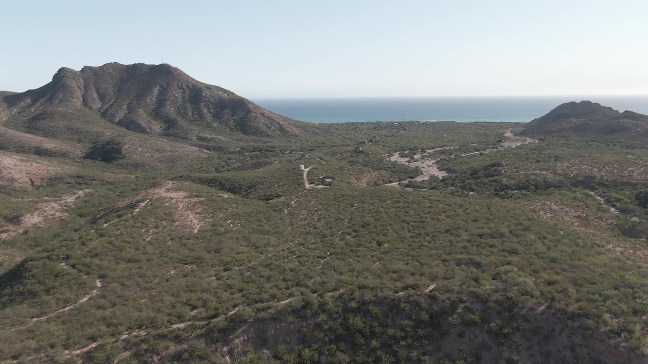 Aerial frame over Cabo Pulmo green scrubland with a large rocky mountain on the left, smaller hill on the right, distant ocean on the horizon, a small structure near the lower center, and no visible people.