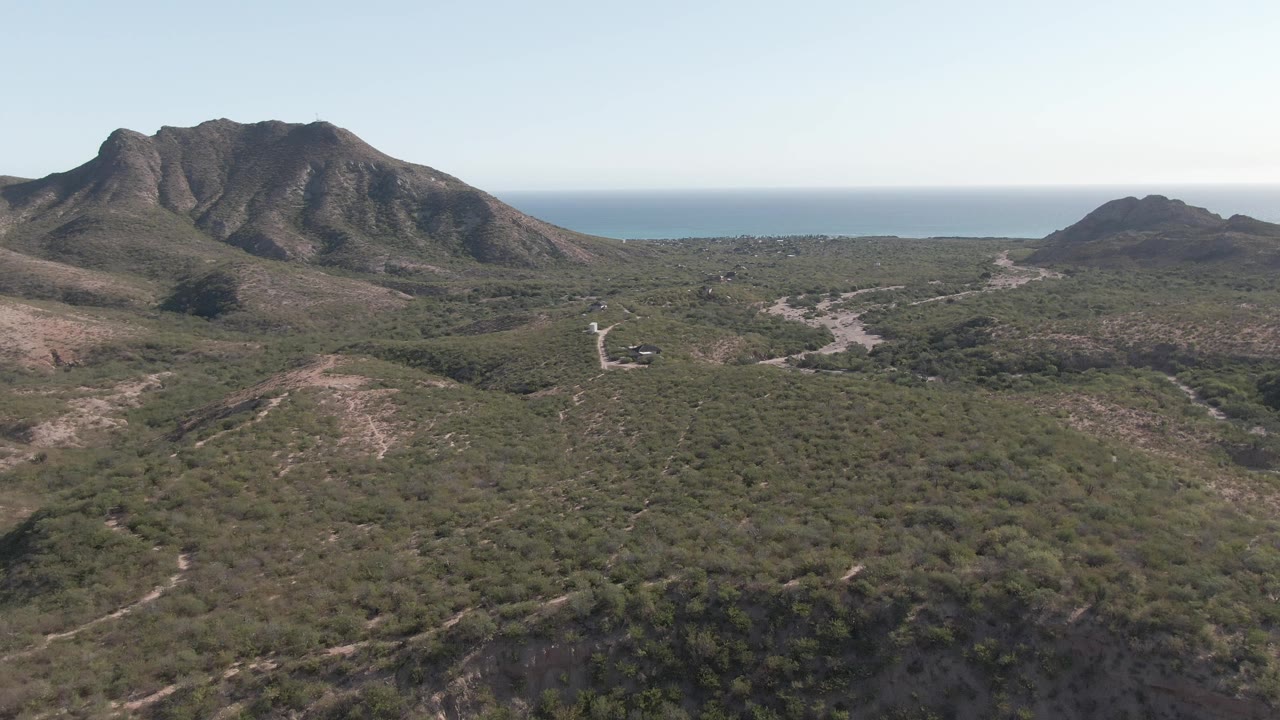 Cabo Pulmo aerial frame with rolling green vegetation, dry wash lines across the midground, a dark rocky mountain ridge at left, a smaller hill at right, distant ocean, and no visible people.