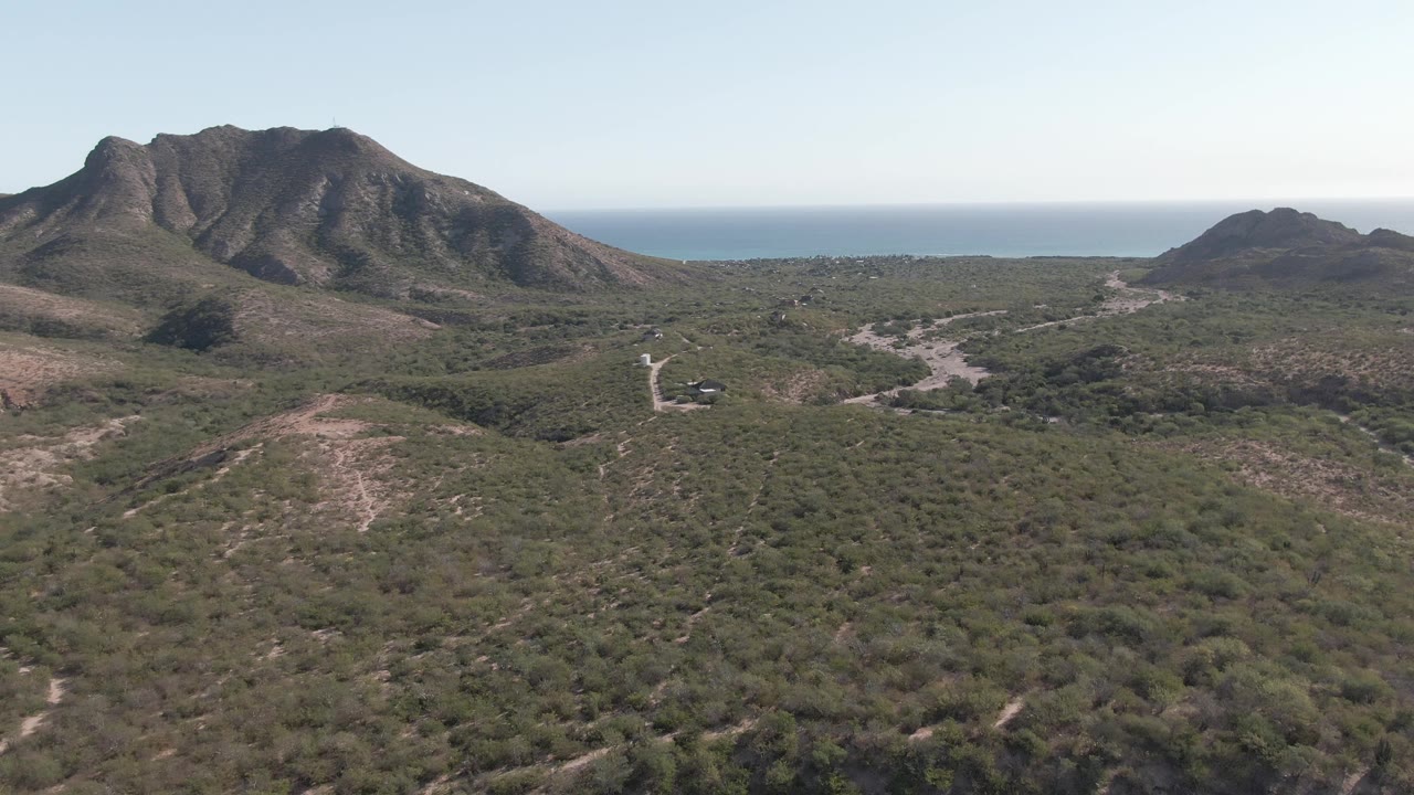 Opening aerial frame over Cabo Pulmo scrubland, with dry hills in the foreground, dark mountain ridges ahead, a blue ocean band beyond, and no visible people.