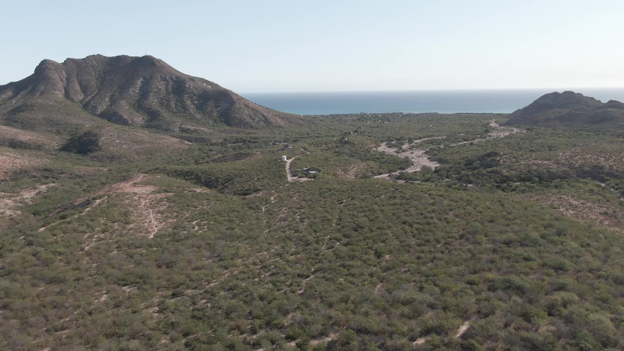 Second aerial frame in Cabo Pulmo showing a broad scrub valley, pale dirt paths, rugged hill faces, open sky, distant ocean, and no visible people.