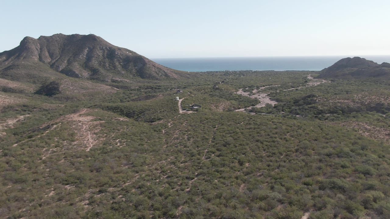Mid-clip aerial frame over Cabo Pulmo with mottled green and brown vegetation in front, a dark rocky ridge near center, a smaller hill to the right, ocean horizon, and no visible people.
