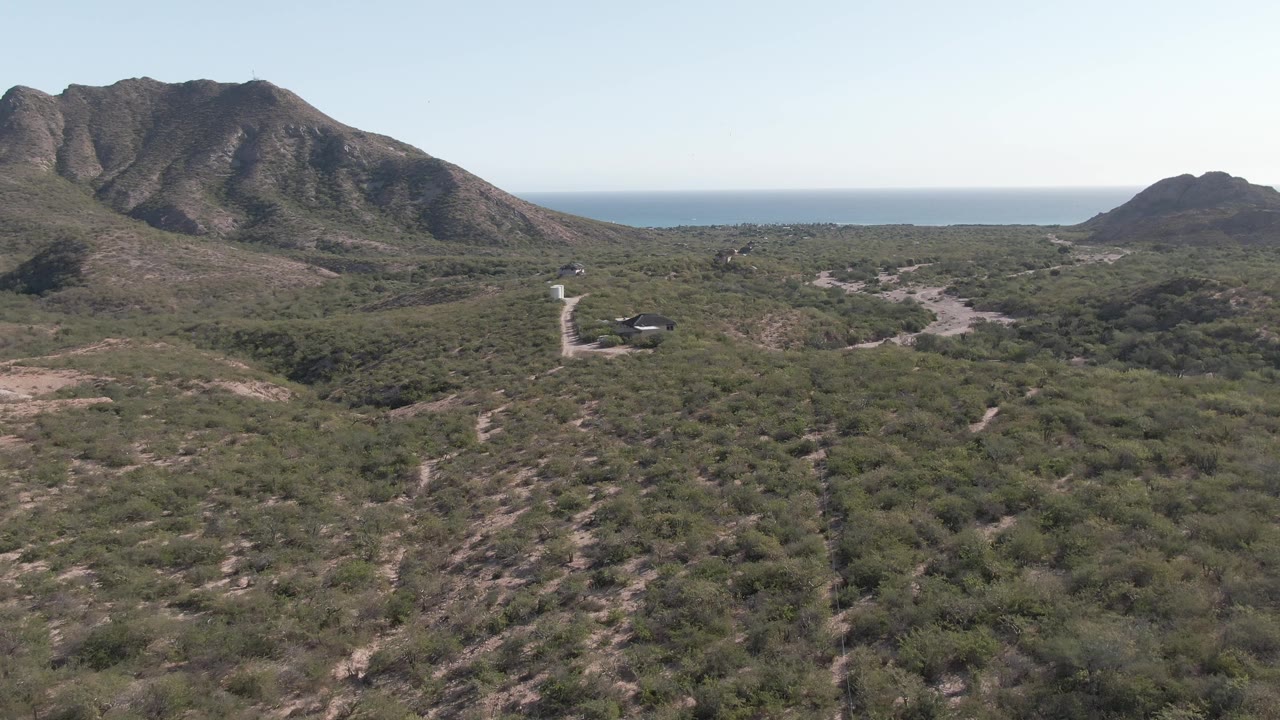 Aerial frame from Cabo Pulmo, Baja California Sur showing scrub-covered terrain, a pale road, rugged mountains, distant blue ocean, and no visible people.