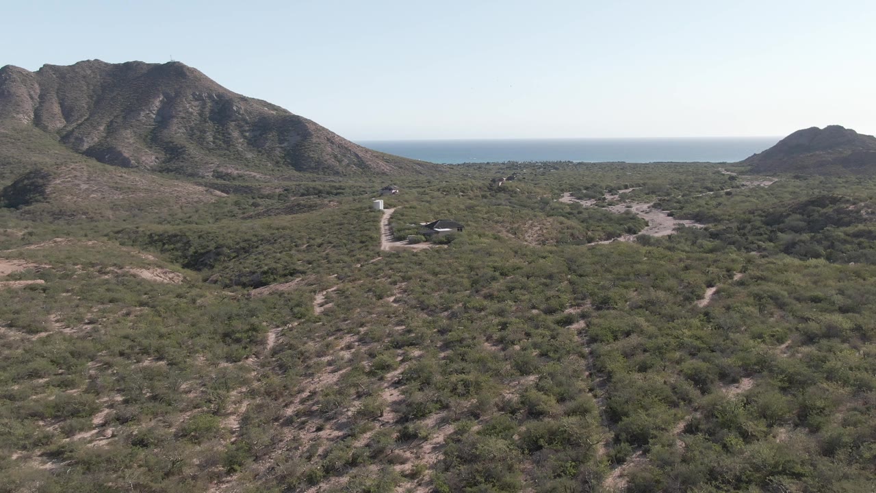 Cabo Pulmo aerial view with the road more central in the scrubland, dark mountain ridges behind it, blue ocean beyond, and no visible people.