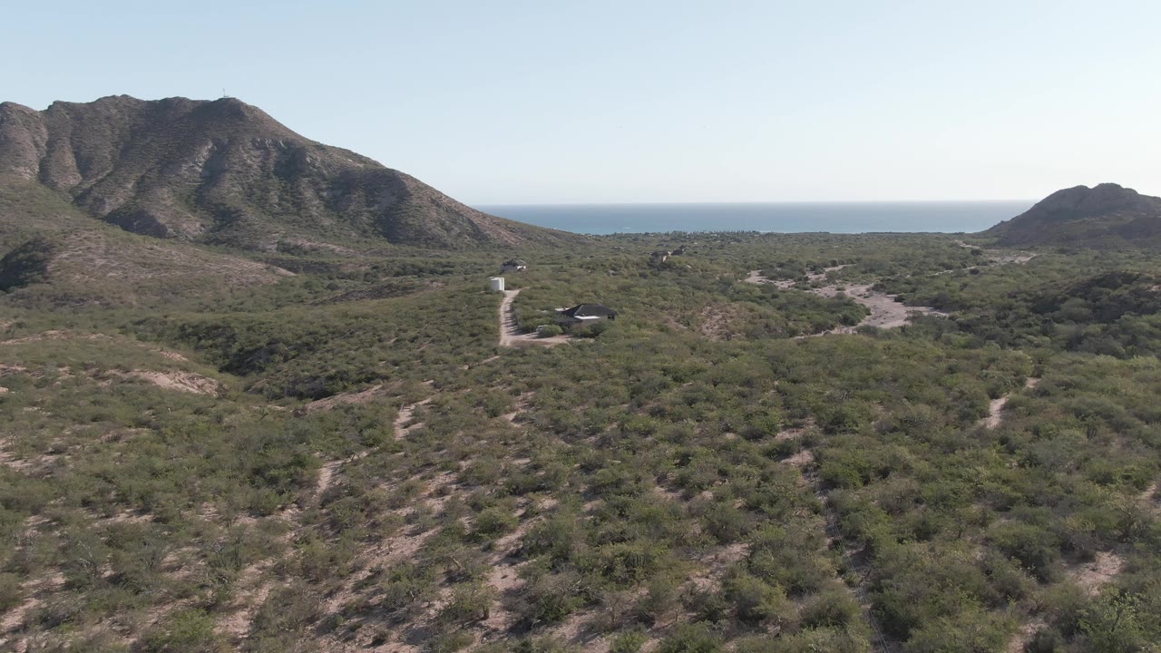Aerial frame in Cabo Pulmo showing the winding pale road and small vehicle below, with scrubland, rocky mountains, distant ocean, and no visible people.